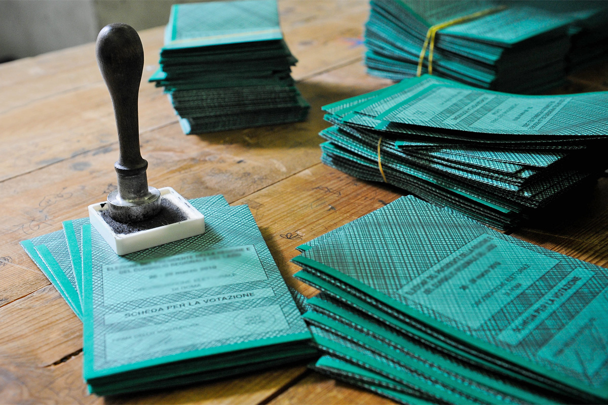 Tessere elettorali di colore verde sul tavolo, pronte per essere vidimate dagli scrutatori con apposito timbro appoggiato sulle tessere.